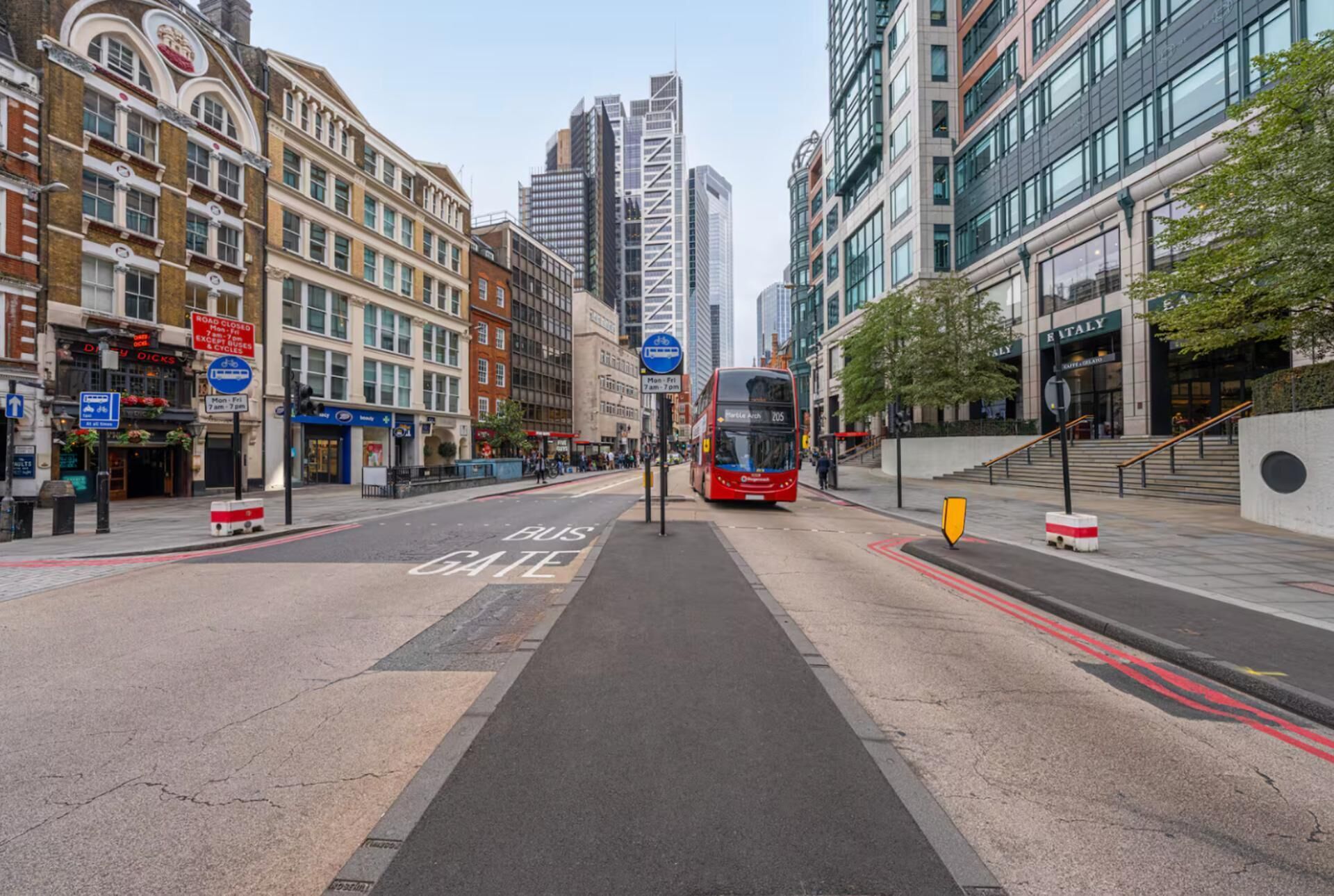 Una calle urbana con un carril para autobuses, edificios y un autobús rojo de dos pisos.