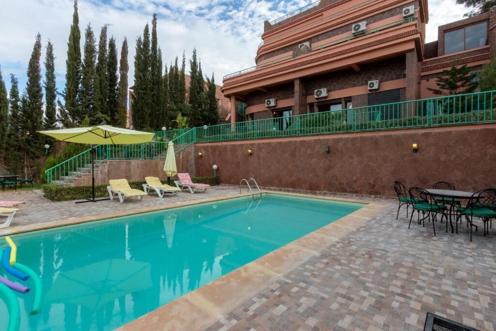 Une piscine entourée de chaises longues et d’une table, le tout bordé par un bâtiment doté d’une rambarde verte et avec des arbres en arrière-plan.