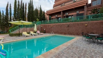 Une piscine entourée de chaises longues et d’une table, le tout bordé par un bâtiment doté d’une rambarde verte et avec des arbres en arrière-plan.