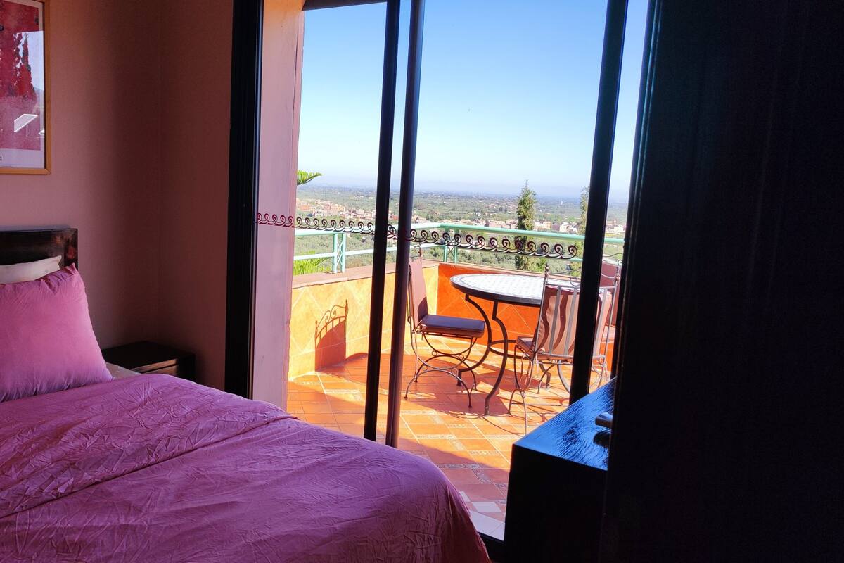 A bedroom with a bed, a framed picture on the wall, and a balcony with a table and chairs.