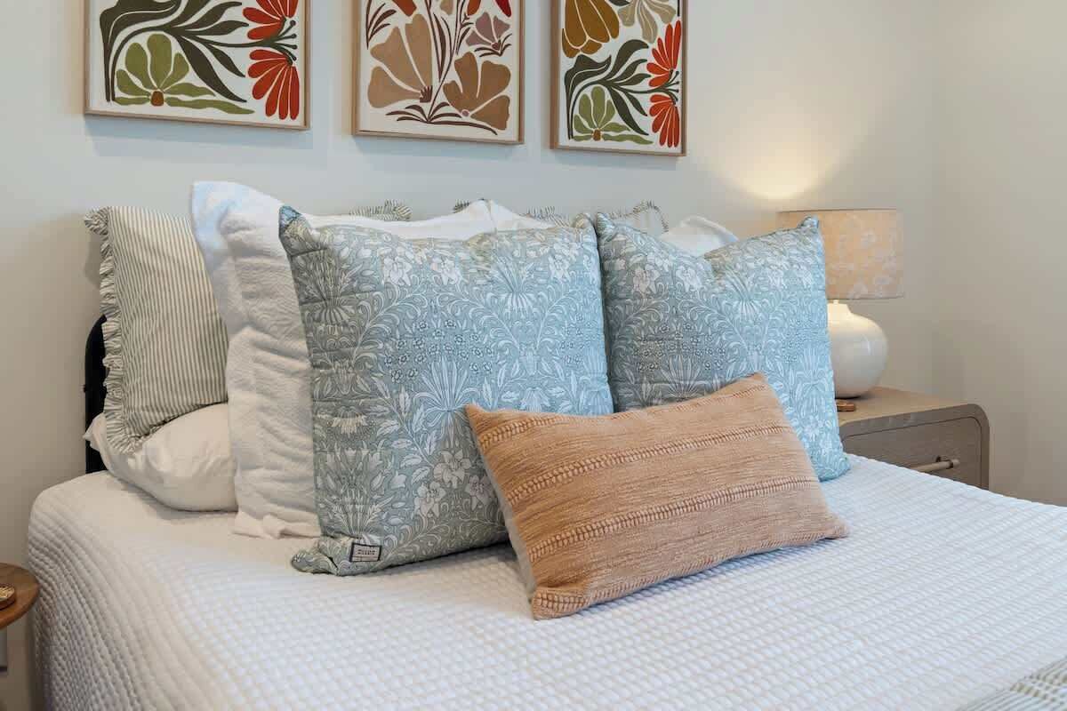 A neatly made bed with patterned pillows and a striped pillow, a bedside lamp, and floral artwork on the wall.