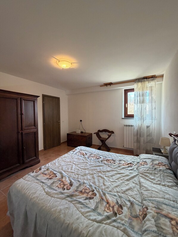 Un dormitorio con cama, armario, mesita de noche, una silla, ventana con cortinas y radiador.