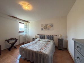 Quarto com cama, mesas de cabeceira, radiador, janela com cortinas, cadeira de madeira e obra de arte na parede.