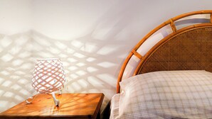 Un letto in vimini con un cuscino a quadretti, un comodino in legno e una lampada intrecciata che proietta ombre sulla parete.