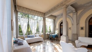 Una habitación con una cama, un sofá, una mesa de comedor y sillas. Hay ventanas con cortinas, una chimenea y un techo con columnas.