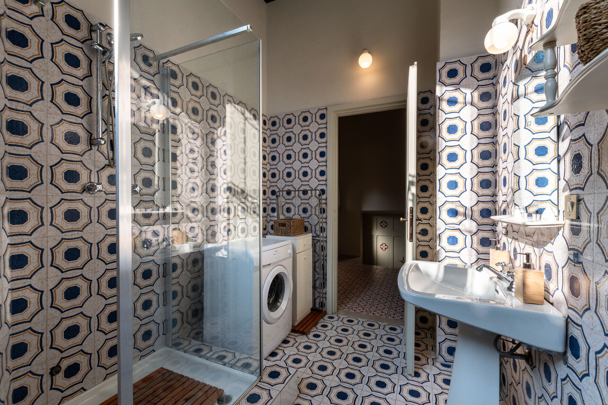 A bathroom with a patterned floor, a washing machine, a glass shower enclosure, and a sink with a mirror.