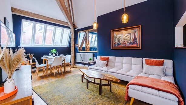 A modern living room with a white sofa, a dining area with a table and chairs, and a large window with a view of the outside. - Stylish 1BDR Duplex Loft Piccadilly (Manchester)