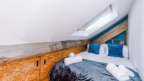 Ein Schlafzimmer mit schrägverdeckter Decke, Holzvertäfelung, einem Dachfenster und einem Bett mit blau-weißer Bettwäsche.