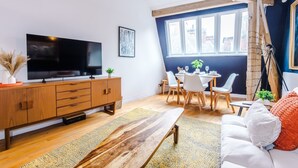 Ein modernes Wohnzimmer mit einem Flachbildfernseher, Holzmöbeln, einem Essbereich mit weißen Stühlen und einem großen Fenster mit Ausblick.