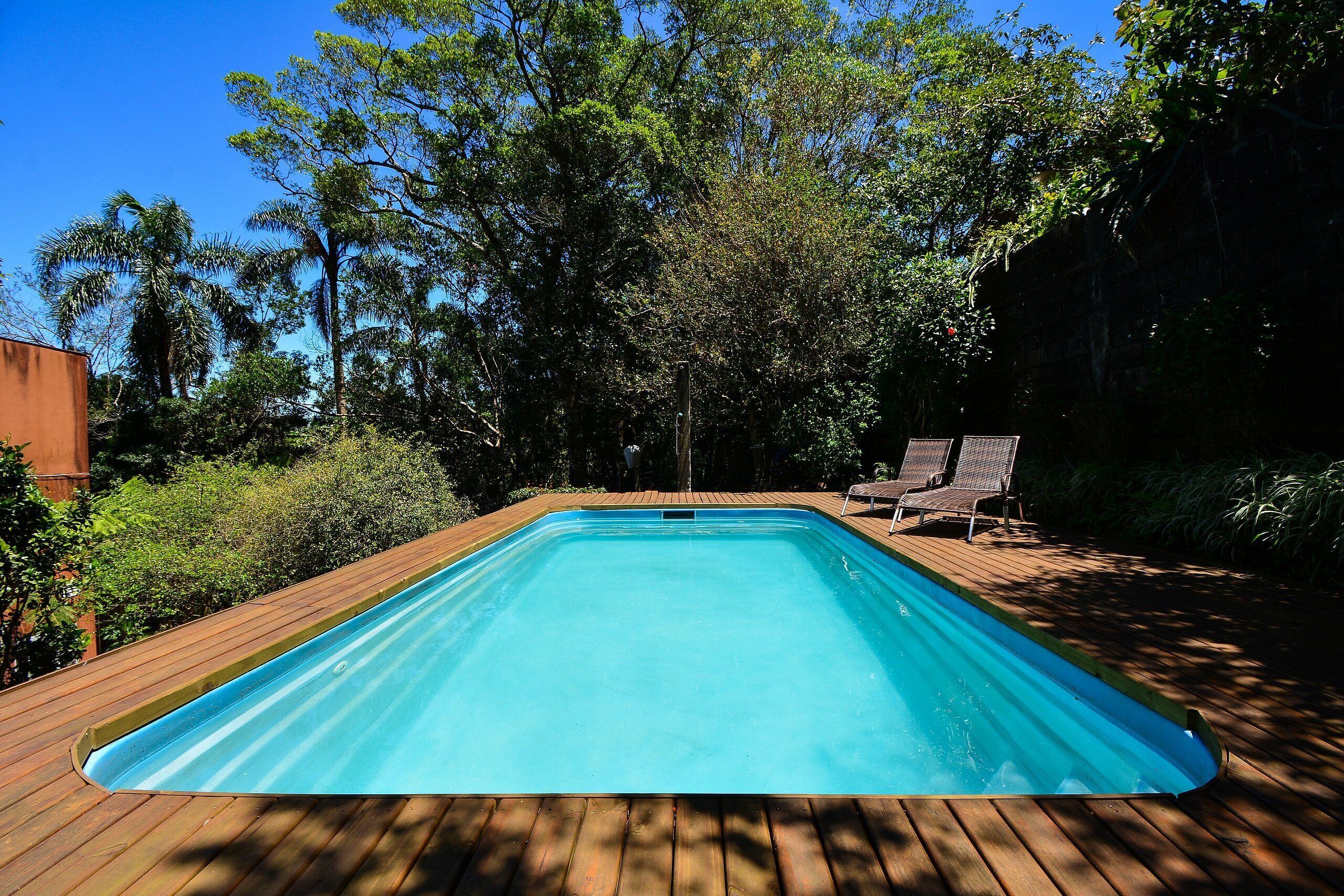 A rectangular swimming pool with a wooden deck, surrounded by trees and a clear blue sky.