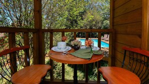A wooden deck with a table set for two, overlooking a swimming pool and lush greenery. - Aconchegante Cabana em Sambaqui - NDI013 (Florianópolis)