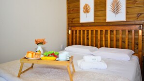 A hotel room with a wooden headboard, a bed with white linens, a bedside table with breakfast items, and two framed leaf artworks on the wall. - Aconchegante Cabana em Sambaqui - NDI013 (Florianópolis)
