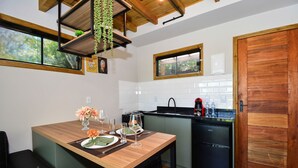 A compact kitchen with a wooden table, a black refrigerator, and a window with a view of greenery. - Aconchegante Cabana em Sambaqui - NDI013 (Florianópolis)