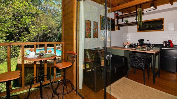 A wooden deck with a table and chairs, a glass door leading to a kitchen, and a pool visible through the door. - Aconchegante Cabana em Sambaqui - NDI013 (Florianópolis)