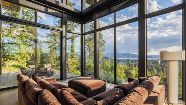 A modern interior with large windows, a brown sectional sofa, and a view of a forested landscape. - The Edge - Modern Retreat with Panoramic Views (Sandpoint)
