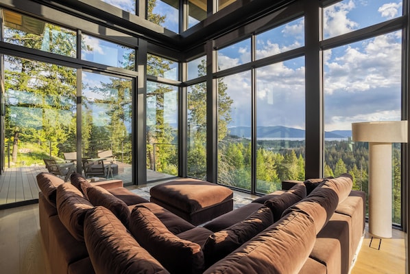 A modern interior with large windows, a brown sectional sofa, and a view of a forested landscape.
