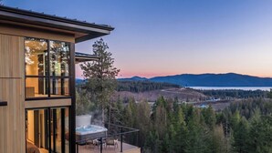 A modern two-story building with large windows, a hot tub on the balcony, and a view of a forested valley. - The Edge - Modern Retreat with Panoramic Views (Sandpoint)