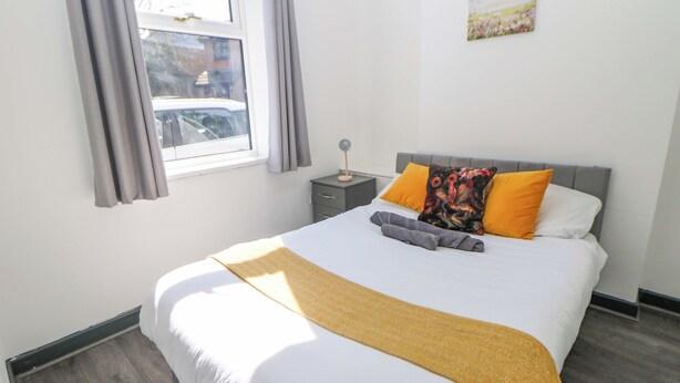 A neatly made bed with a white comforter and a yellow throw blanket, a gray headboard, and two pillows, one with a floral pattern. - Potter's Wheel (Stoke-on-Trent)