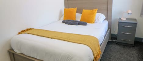 A neatly made bed with a headboard, a bedside table with a lamp, and a small drawer unit.