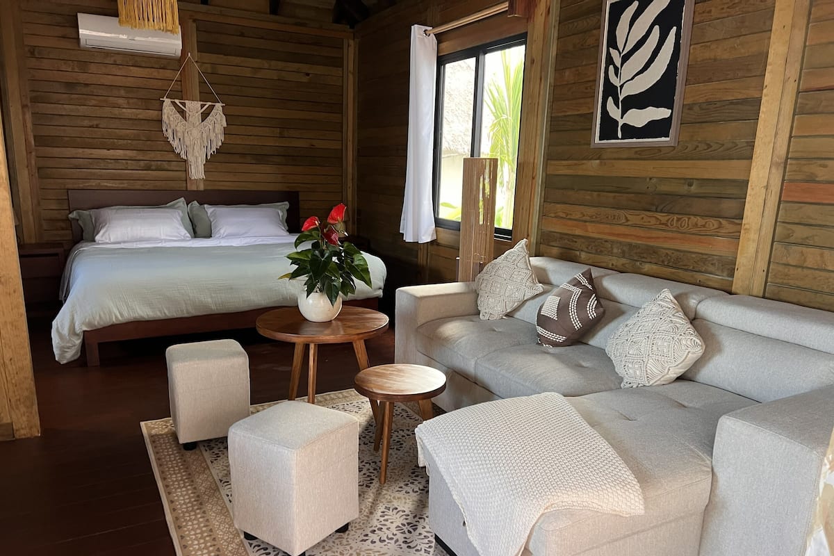 A wooden cabin interior with a bed, sofa, ottoman, and a small table with a plant.