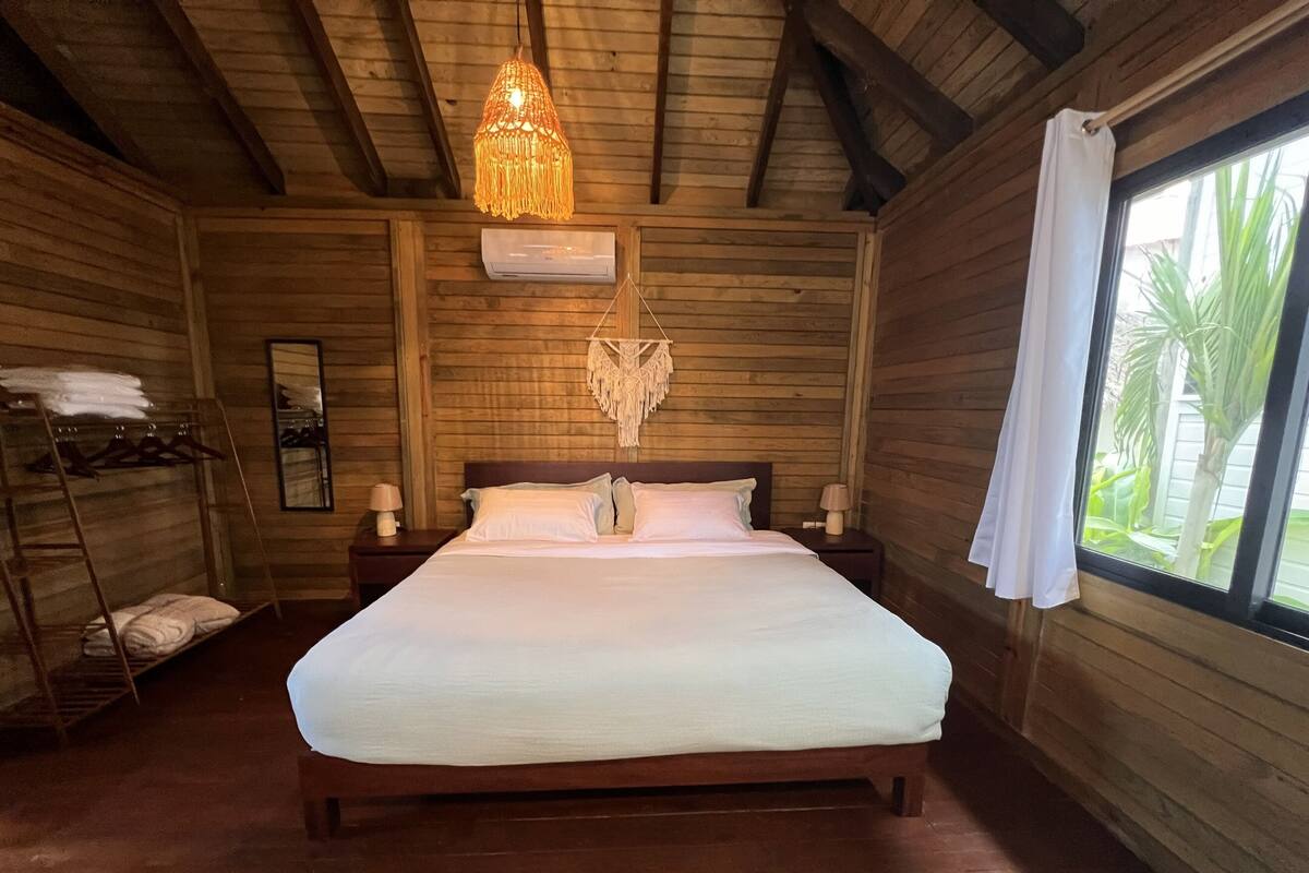 A wooden room with a bed, a hanging light fixture, a macrame wall hanging, and a window with a view of greenery.
