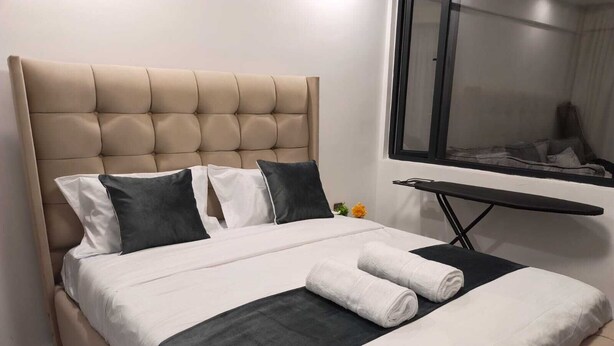 A bed with a tufted headboard, white and dark pillows, a black bed runner, and a rolled white towel. - Charming 1-bedroom apartment in vibrant Nairobi with fitness room (Nairobi)