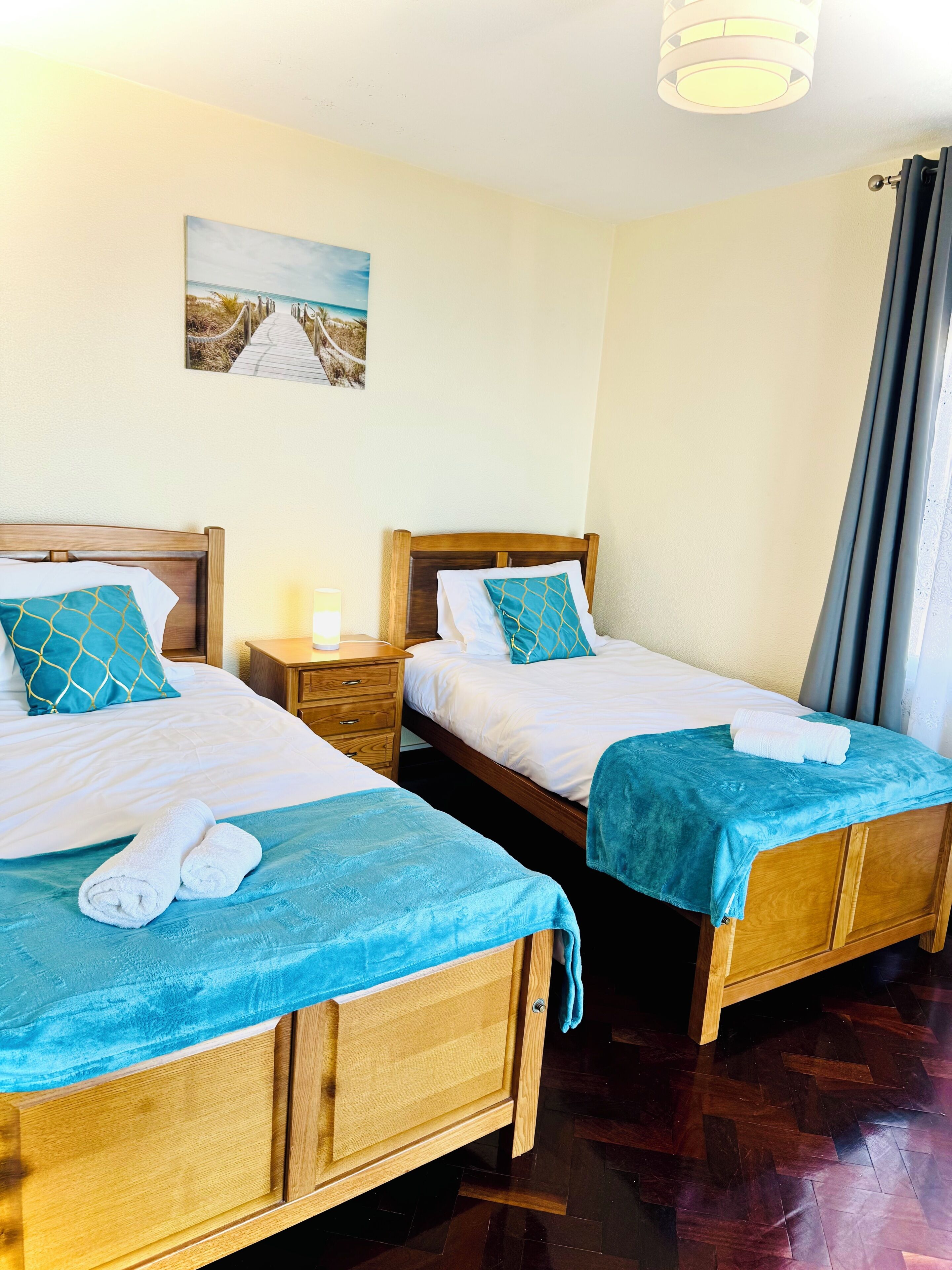 A hotel room with two wooden beds, each with a blue bedspread and matching pillows, a nightstand, a painting on the wall, and a window with curtains.