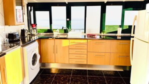 Cozinha compacta com máquina de lavar, cafeteira e vista para o mar através de grandes janelas.