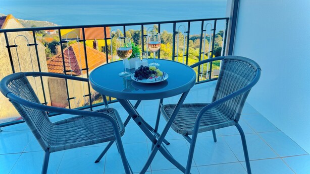 Varanda com mesa e cadeiras, um prato de uvas e uma taça de vinho, e vista para o mar.