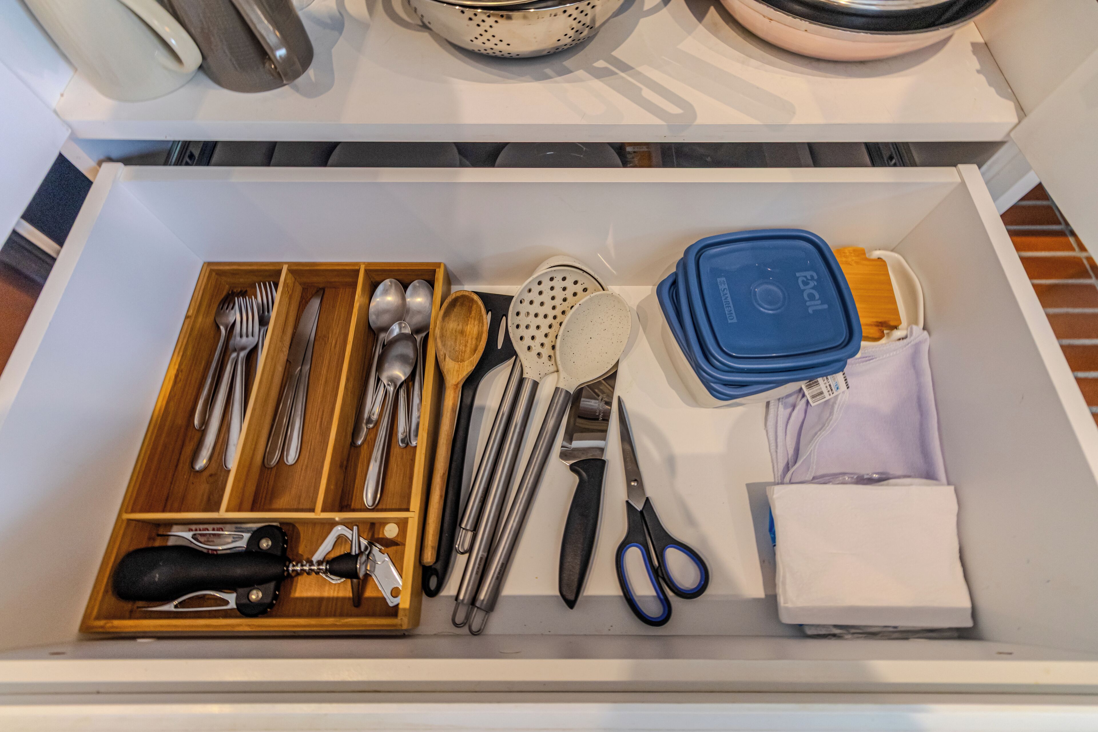 A drawer with utensils, a colander, a pair of scissors, and a wooden utensil box.