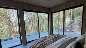 Une chambre avec un grand lit, un plafond en bois et une vue sur une forêt grâce à de grandes fenêtres du sol au plafond.