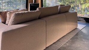 Un salon moderne doté d’un grand canapé d’angle beige, d’une cheminée et de grandes fenêtres donnant sur une forêt.