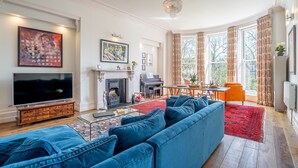 A living room with a large TV, blue sofa, fireplace, dining area, and large windows with curtains. - The Classic on the Mount -  a premium stay that sleeps 4 guests  in 2 bedrooms (York)