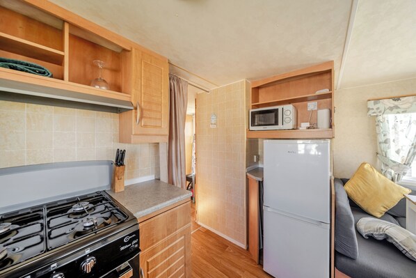 Une cuisine compacte équipée d’une cuisinière à gaz, d’un four à micro-ondes et d’un réfrigérateur, de placards en bois et d’un petit coin salon avec des coussins.