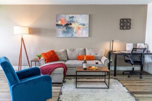Una sala de estar con un sofá, una mesa de centro, un sillón azul, una lámpara y un cuadro en la pared.