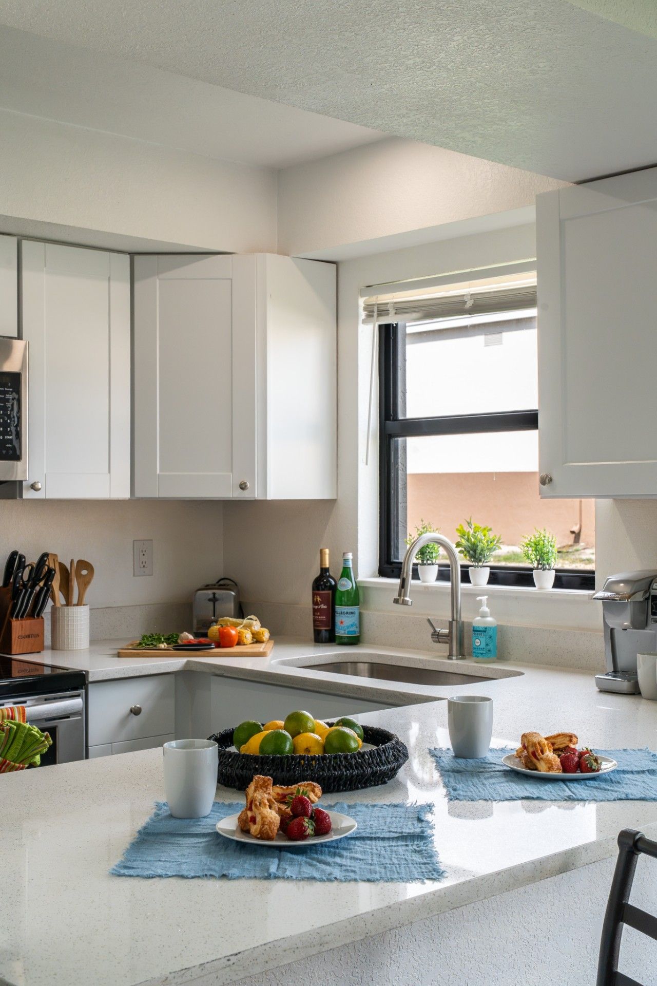Una cocina moderna con armarios blancos, horno empotrado, lavamanos y ventana con plantas en macetas.
