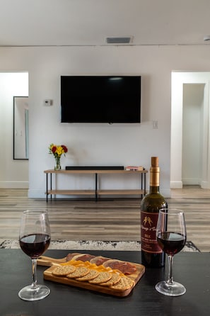 Habitación con una televisión de pantalla plana montada en la pared, un banco de madera y una mesa con una botella de vino, copas y una tabla de queso y galletas.