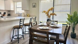 A dining area with a table set for four, surrounded by wooden chairs, a kitchen area with white cabinets, and a window with blinds. - Modern Coral Gem: Patio | Work Space | Downtown (Cape Coral)