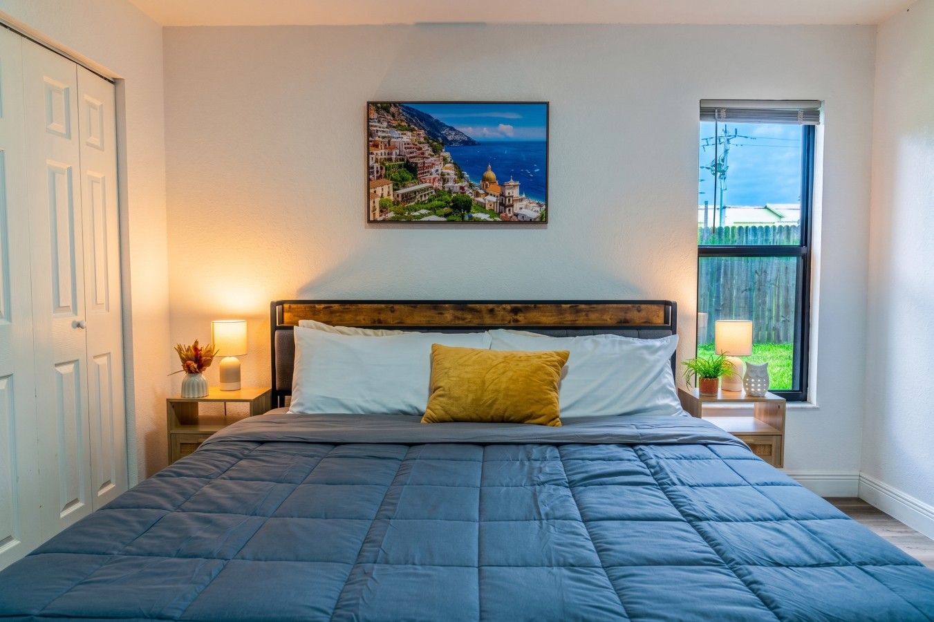 A bedroom with a bed, a nightstand, a lamp, a window with a view, and a painting on the wall.