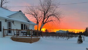 Ein Haus mit einer Holzterrasse, einem mit Schnee bedeckten Garten und einem Sonnenuntergang im Hintergrund.