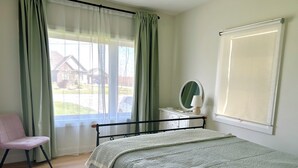 Ein Schlafzimmer mit einem Bett, einem Sessel, einer Kommode mit Spiegel und einem Fenster mit Vorhängen.