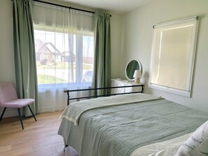 Une chambre à coucher avec un lit, une chaise, une commode avec un miroir et une fenêtre avec des rideaux.