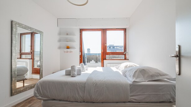 Un dormitorio con una cama, un espejo grande, una ventana y vista a un edificio.