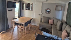 A living room with a wooden table, chairs, a sofa, a TV, a ceiling fan, and a wall-mounted air conditioner. - Mobile home near the sea, Camping 4* Mar Estang (Canet-en-Roussillon)