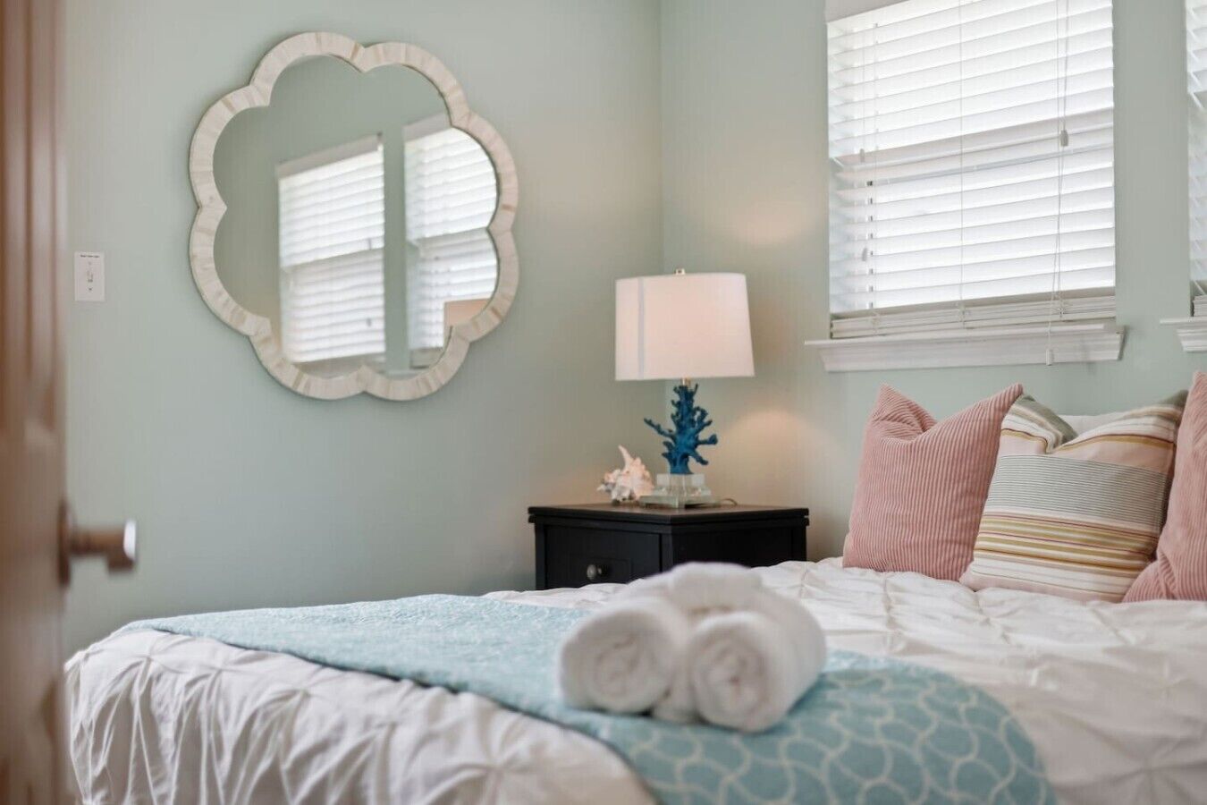 Un dormitorio con una cama, una mesita de noche con lámpara, un espejo y una ventana con persianas.