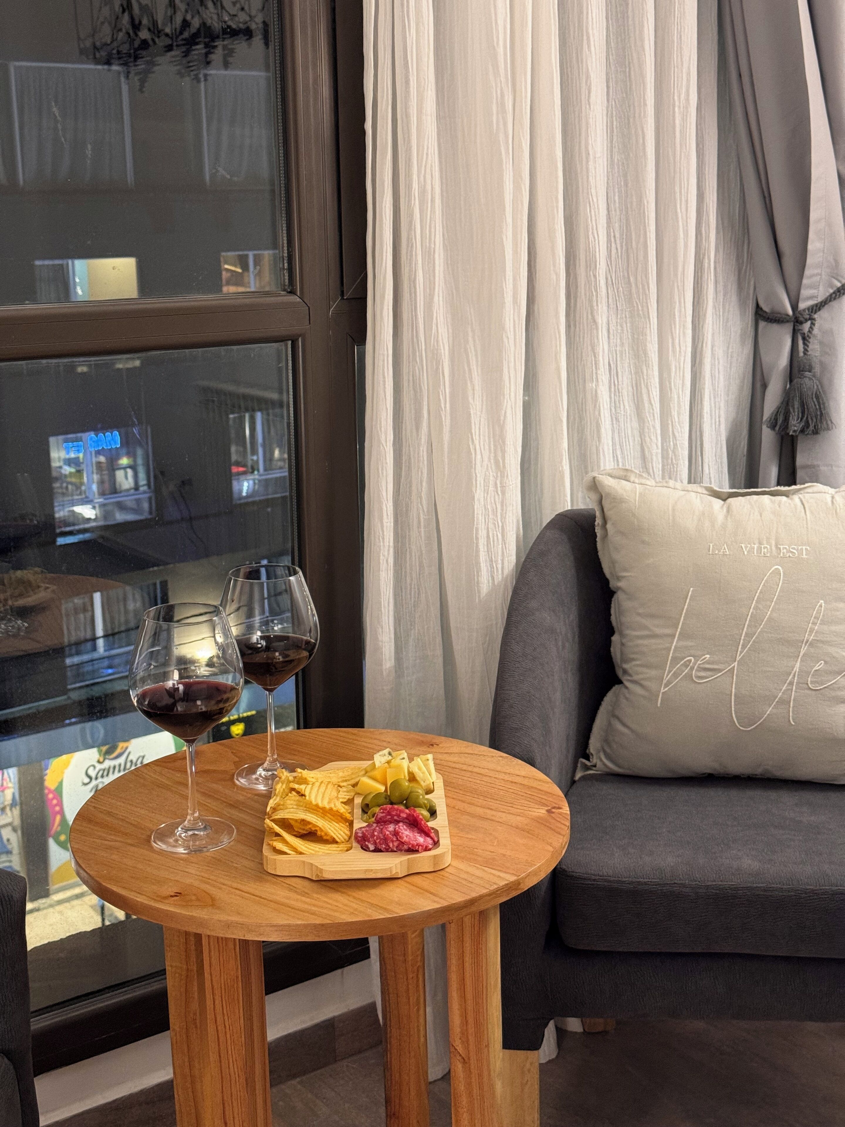 A cozy seating area with a round wooden table, two wine glasses, and a cheese board with various cheeses and olives.