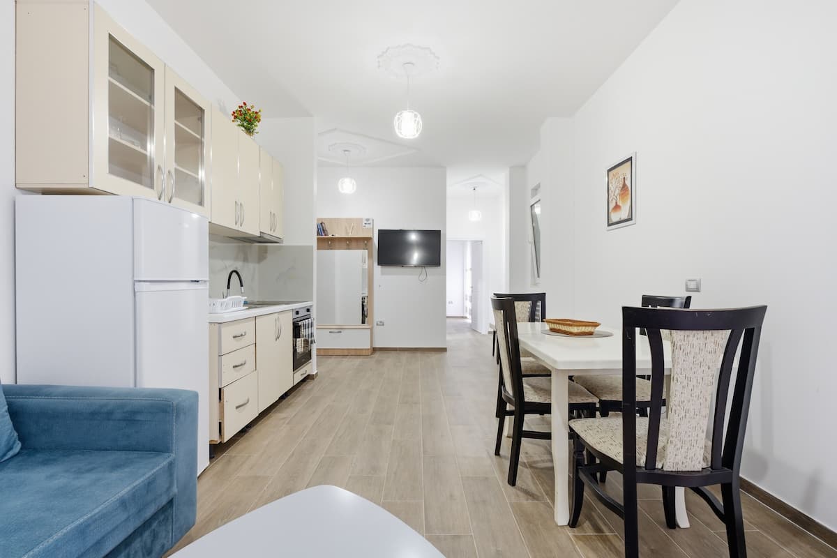 Una cocina moderna con armarios blancos, un comedor con mesa y sillas, y un sofá azul.