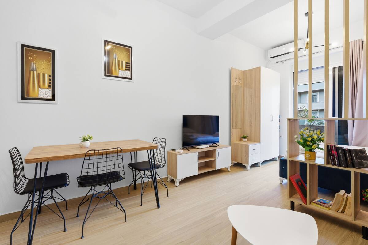 Una sala de estar moderna con una mesa de comedor de madera, sillas de estructura metálica negra, un televisor, una estantería y un ventanal grande.