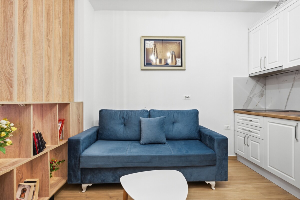 A living room with a blue sofa, a white chair, a wooden bookshelf, and a kitchen area with white cabinets and a marble countertop.