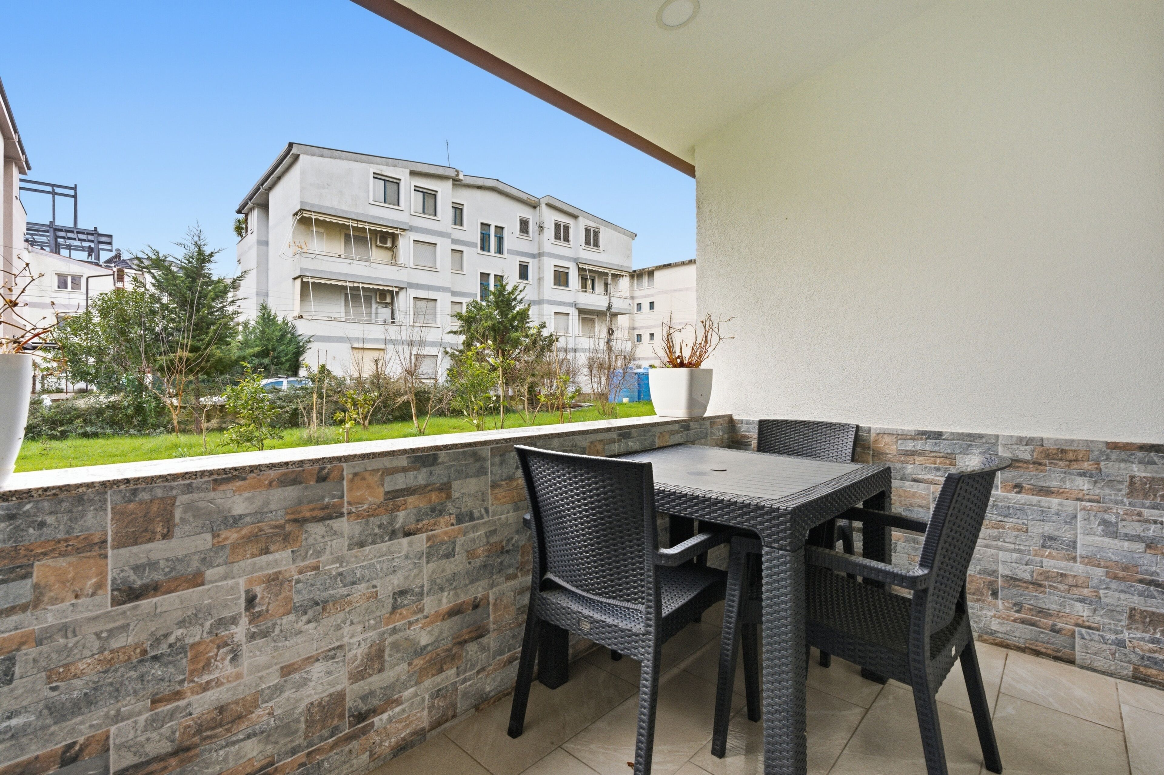 A balcony with a table and chairs, a stone wall, and a view of a multi-story building.